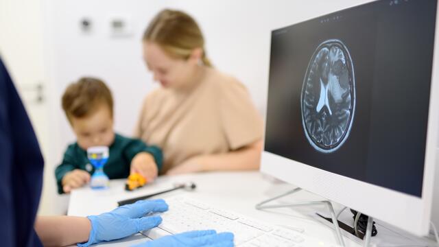 Parent and child meeting with a pediatric neurologist sitting at a desk with a brain scan on their computer